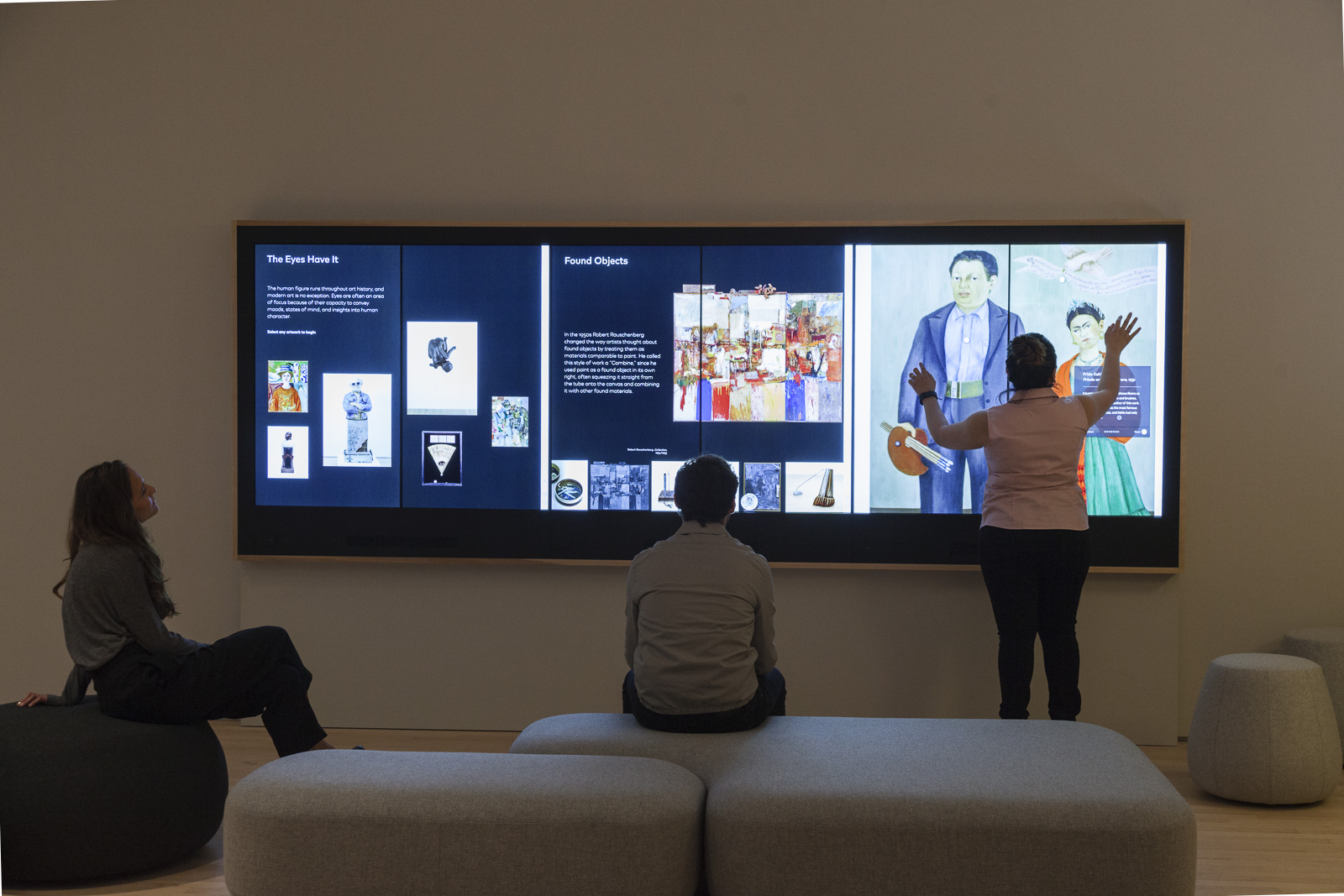 A photo inside SFMOMA's Painting an Sculpture Interpretive Gallery shows three visitors interacting with the Wall Experience. Two are seated on cushions while a third visitor uses both hands to interact with a large touch screen.