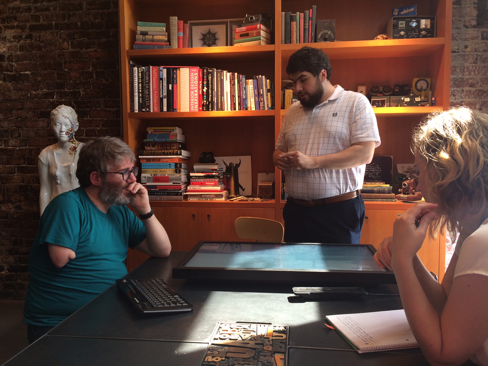A photo inside the Belle & Wissell, Co. studio shows three individuals talking around a touch-screen laid on a conference table. From left to right are Shaun Kane, Sina Bahram, and Sarah Trueblood.